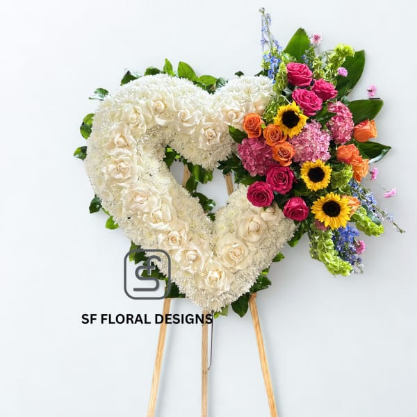 Heart-shaped white standing wreath with colorful roses, sunflowers, and mixed blooms on one side.