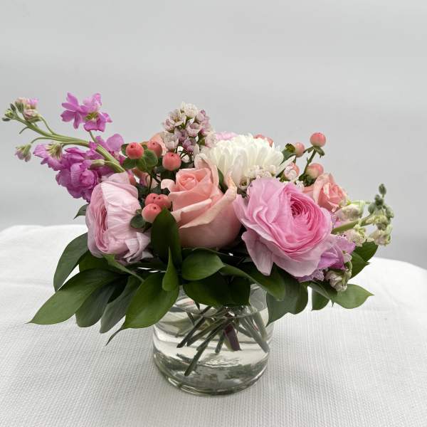 Low arrangement of soft pink and white roses and ranunculus in a clear glass vase.
