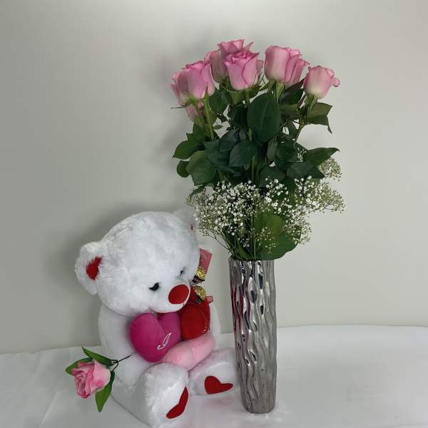 Pink roses in a tall silver vase beside a white teddy bear holding hearts