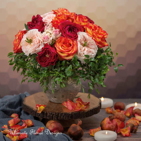 A round arrangement of red, orange, and blush roses in a stone vase.