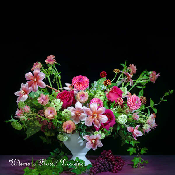Pink and magenta floral arrangement in a white vase with grapes at the base