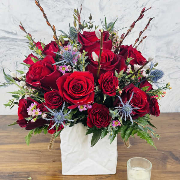 Red rose arrangement in a white square vase with blue thistle accents