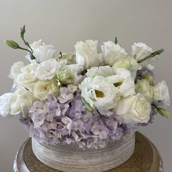 White roses and pale lavender flowers arranged in a low round box.