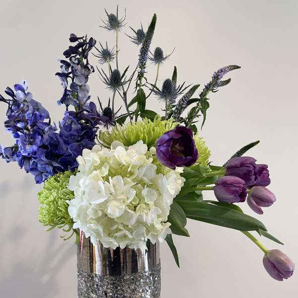 Mixed bouquet with purple tulips, blue delphinium, and white hydrangea in a silver vase