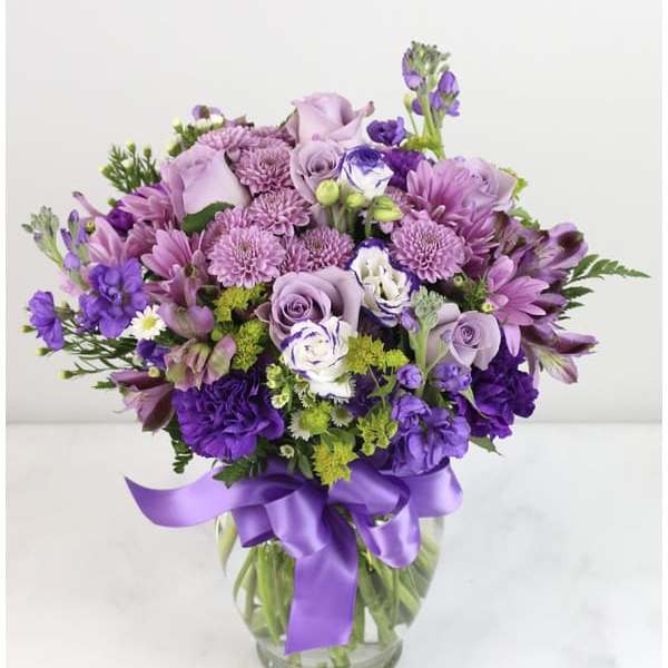 Lavender and purple mixed flower arrangement with roses and mums in a clear vase with a satin ribbon bow