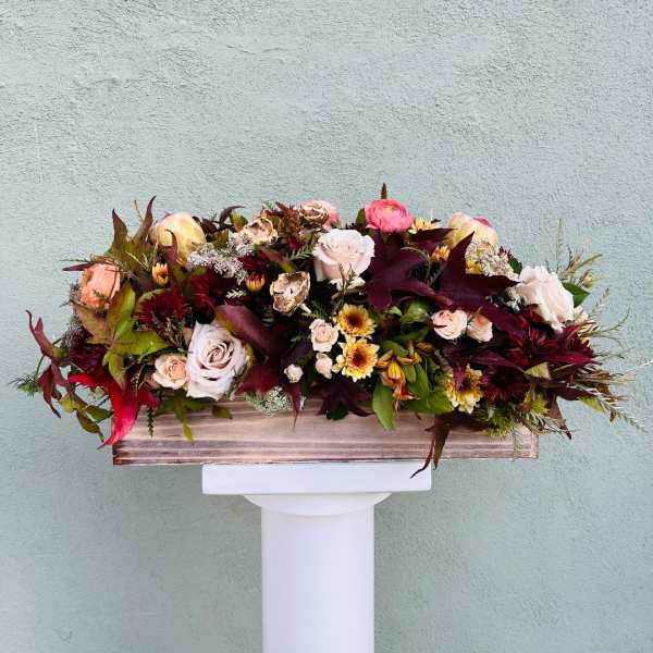 Low rectangular arrangement of blush roses and autumn-toned flowers in a wooden box on a white pedestal.