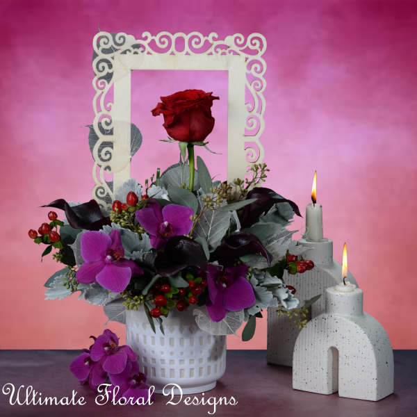 Red rose and purple orchid arrangement in a white vase with candles