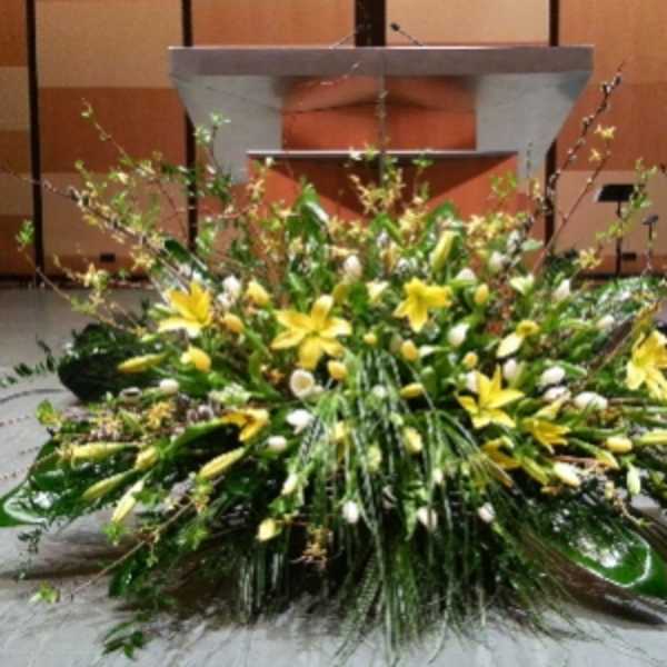 Large low yellow and white lily arrangement with abundant greenery in front of a podium