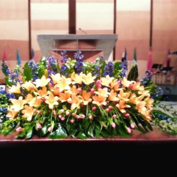Low altar spray of peach lilies, blue flowers, and pink buds in a lush horizontal display.
