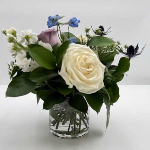 Small arrangement of cream and lavender roses with blue delphinium in a clear glass vase