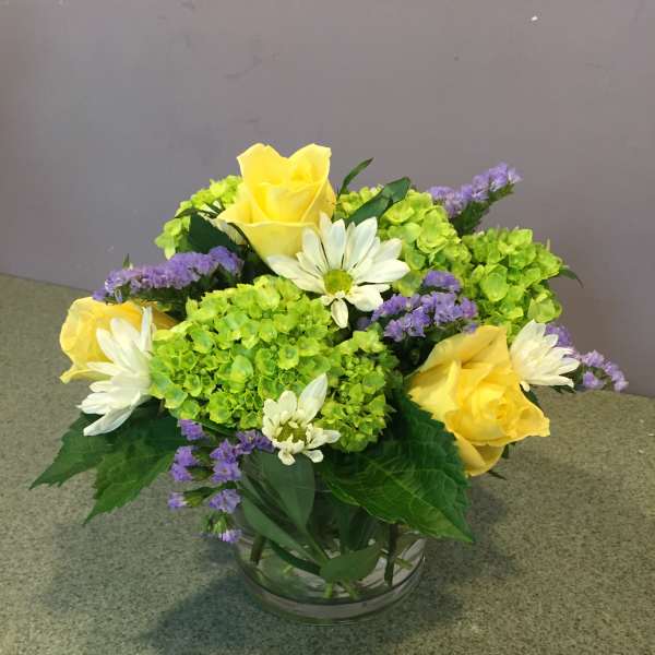 Yellow roses and white daisies in a glass bowl with green hydrangeas and purple filler flowers