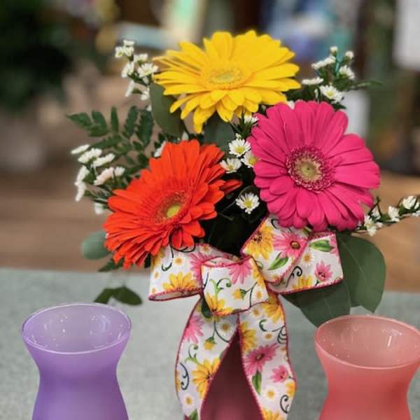 Bright gerbera daisies in a frosted pink vase with floral bow, flanked by two empty frosted vases.