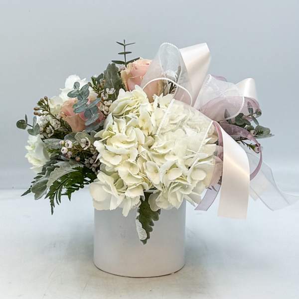 White hydrangeas and blush roses in a white cylinder vase with sheer white and pink ribbon bow