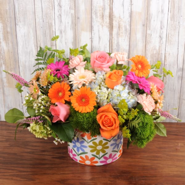 Low arrangement of orange and pink roses and gerbera daisies with blue hydrangea in a colorful round container