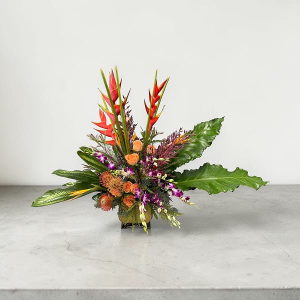 Tropical arrangement with orange roses, protea, purple orchids, and red heliconia in a low glass vase