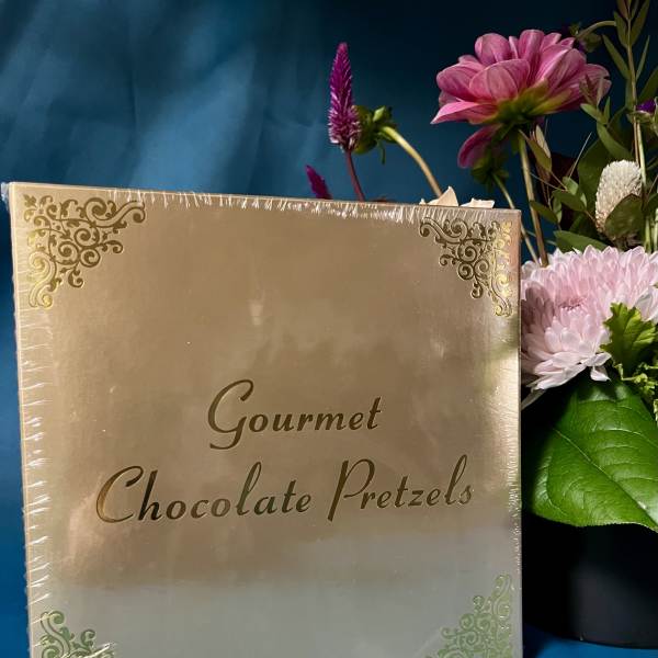 Gold box of gourmet chocolate pretzels beside a pink and purple flower arrangement on a teal background