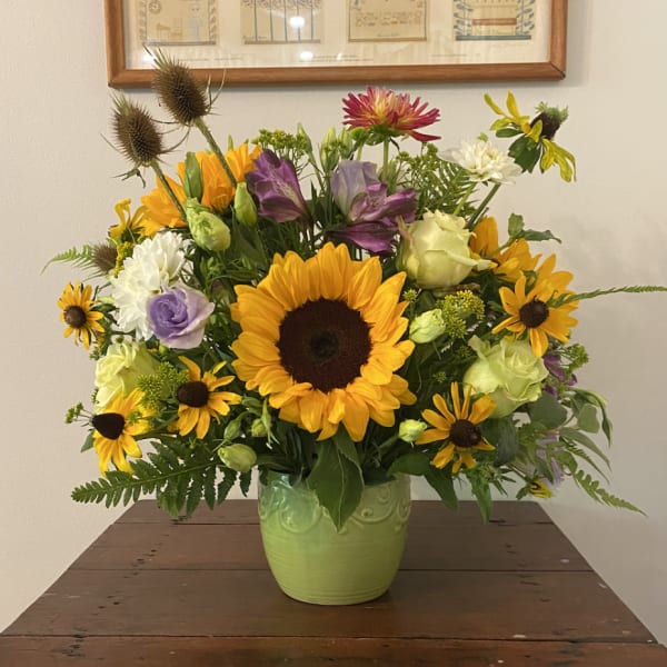 Mixed arrangement with sunflowers, yellow daisies, roses and purple blooms in a green ceramic vase
