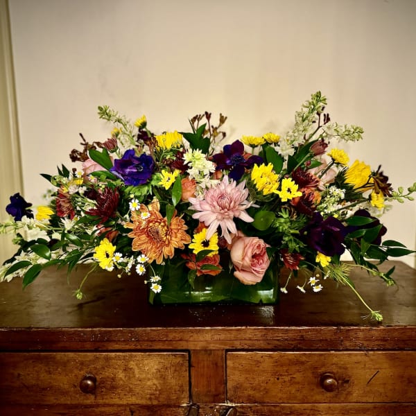 Low rectangular arrangement of mixed yellow, pink, orange, and purple flowers on a wooden dresser