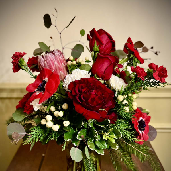 Red and white floral arrangement with roses, anemones, and evergreens in a glass vase on a wooden table.