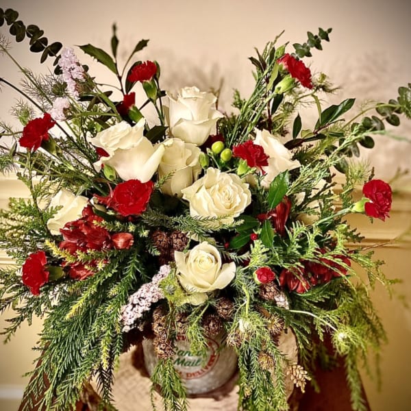 Rustic arrangement of white roses and red carnations with winter greenery in a metal container on a wood slice.