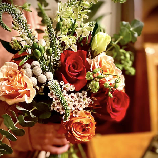 Hand-tied bouquet with red and orange roses, peach blooms, and small white accent flowers.
