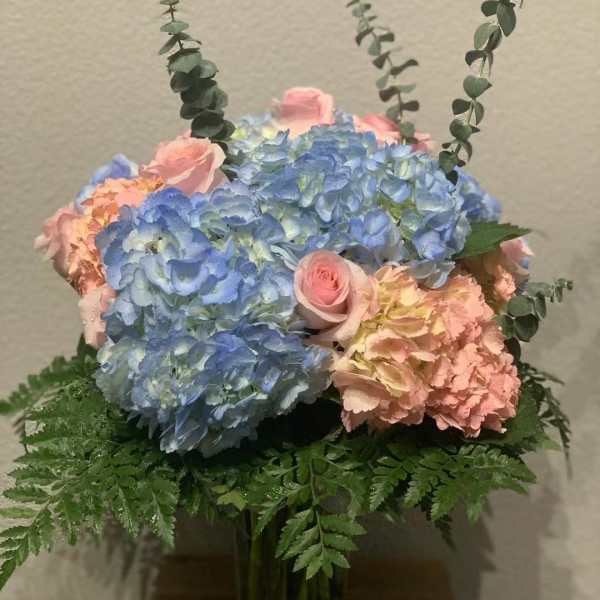 Pastel blue and pink hydrangeas with pink roses and eucalyptus in a clear glass vase with a fern collar