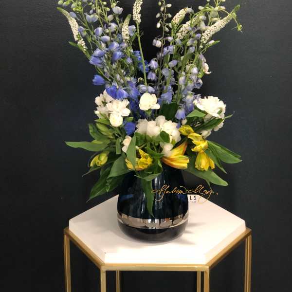 Tall mixed bouquet in a dark glass vase with blue, white, and yellow flowers