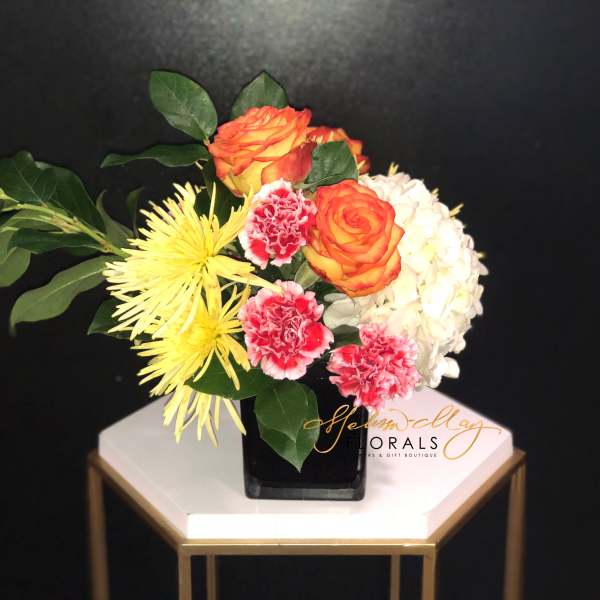 Bouquet of orange roses, yellow spider mums, pink carnations, and white hydrangea in a black vase