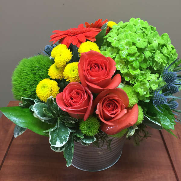 Compact arrangement of red roses, red gerbera, and mixed yellow and green blooms in a metal pot.