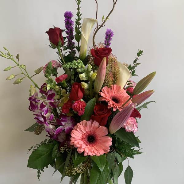 Tall mixed bouquet with pink gerberas, red roses, calla lilies, and orchids in a glass vase