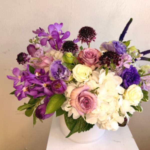 Low vase arrangement of purple orchids, lavender roses, white hydrangeas, and mixed blooms in a white pot