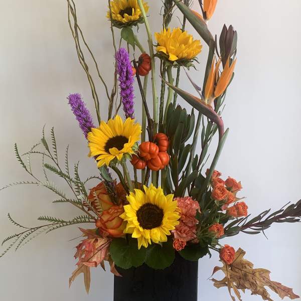 Tall floral arrangement with sunflowers, orange blooms, and purple spikes in a black vase