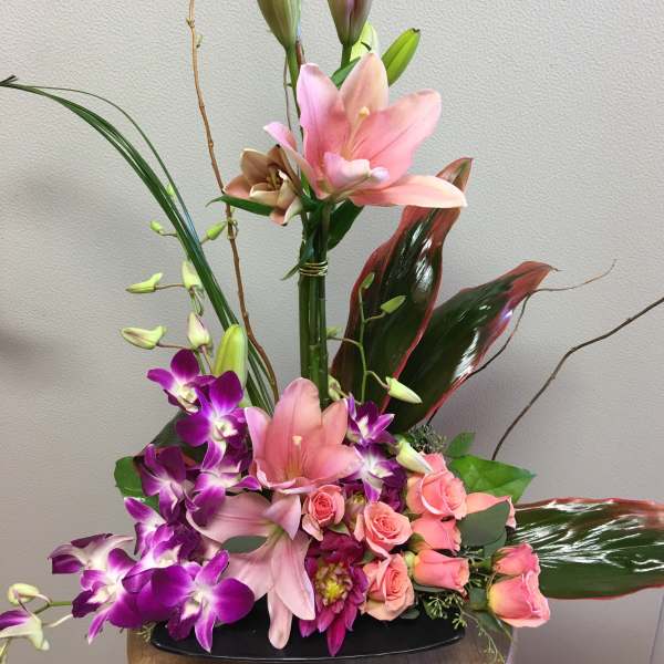 Tall floral arrangement with pink lilies, purple orchids, and roses in a black vase