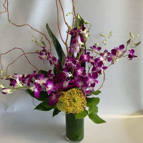 Purple orchids and a yellow pincushion bloom in a tall glass vase.