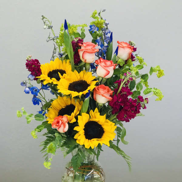 Bouquet of sunflowers, coral roses, and blue flowers in a glass vase
