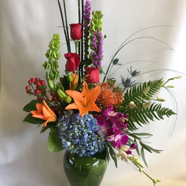 Tall mixed flower arrangement in a green glass vase with lilies, roses, orchids, and hydrangea