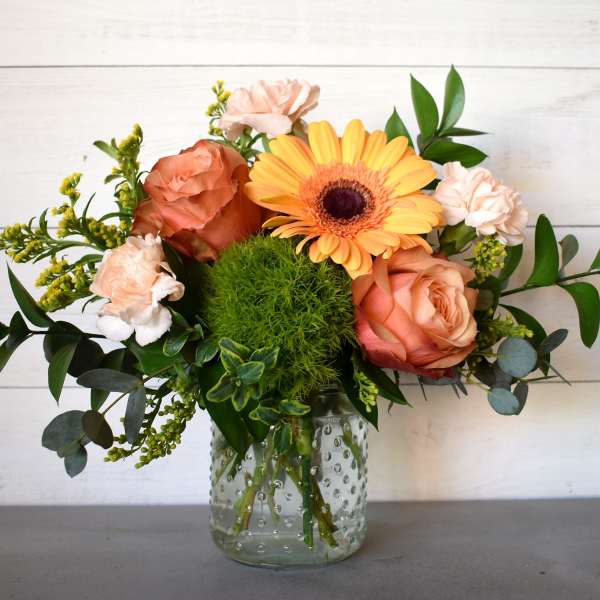 Peach roses, pale carnations, a yellow gerbera daisy, and a green pompom bloom in a clear glass jar.