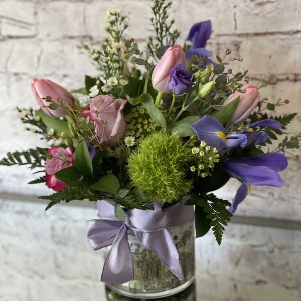 Mixed bouquet of pink tulips, roses, and purple iris in a glass vase with lavender ribbon