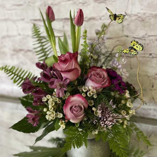 Pink roses, tulips, and mixed blooms in a silver vase with yellow butterfly picks.