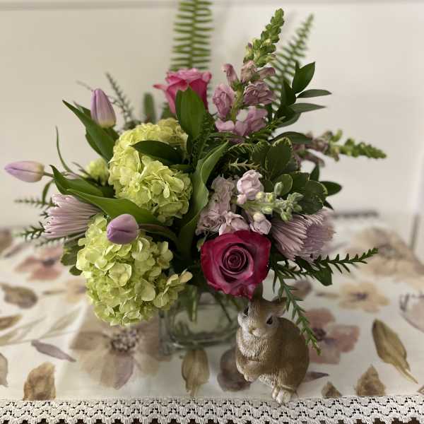 Spring arrangement with green hydrangeas, pink roses, and tulips in a glass vase beside a bunny figurine.