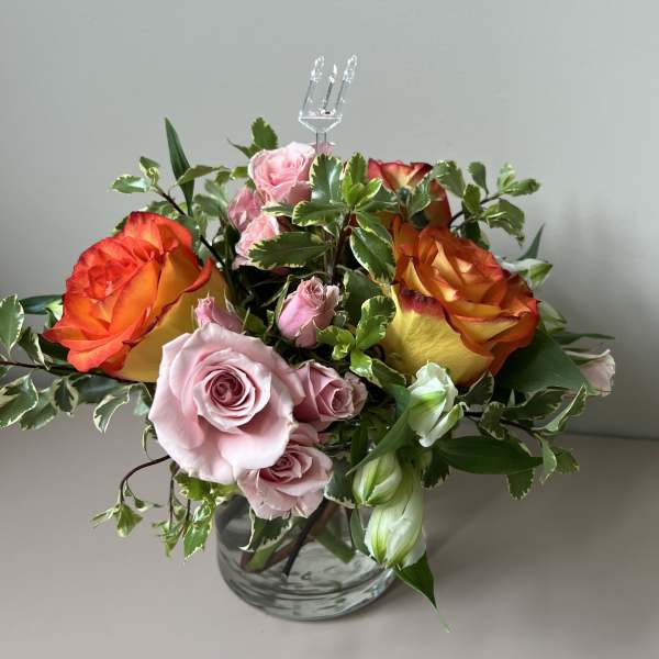 Short glass vase arrangement with orange-yellow and pink roses and variegated foliage