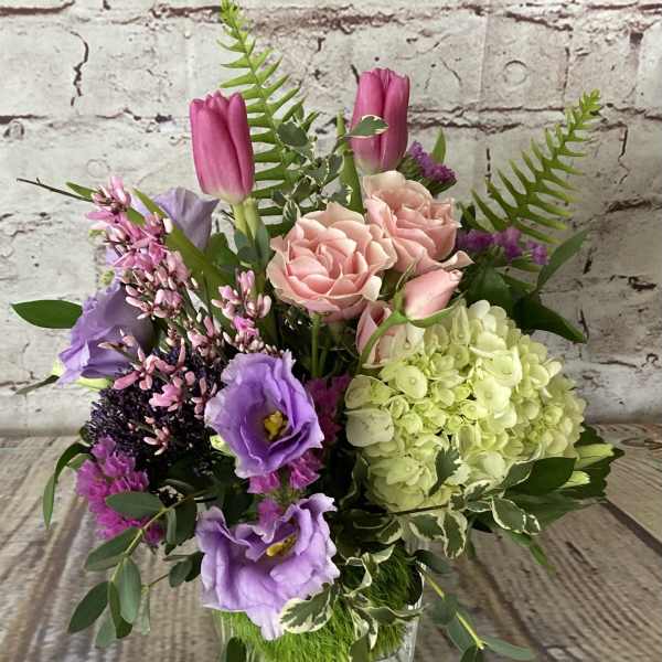 Mixed arrangement of pink roses, purple tulips, lavender lisianthus, and green hydrangea in a glass cube vase