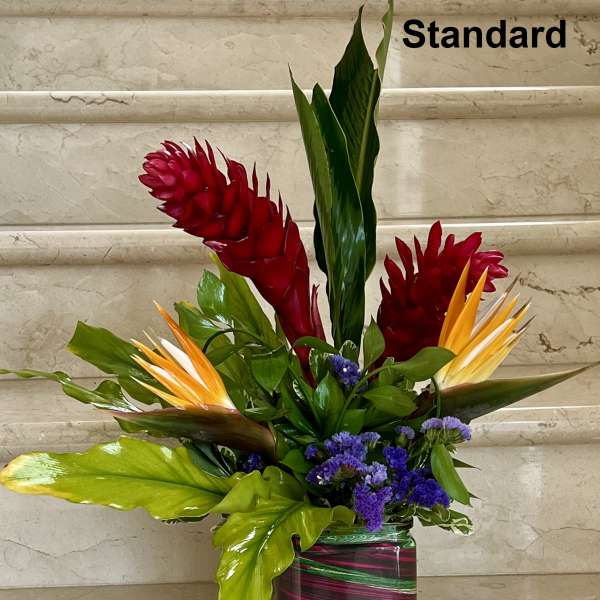 Tall tropical arrangement with red ginger, orange blooms, and purple flowers in a striped square glass vase.