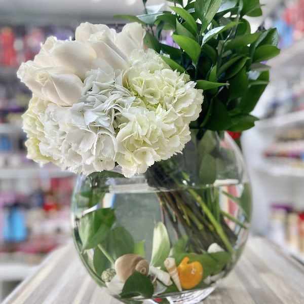 Round glass vase with white roses and hydrangeas, tall greenery, and seashells with a small yellow duck in the water