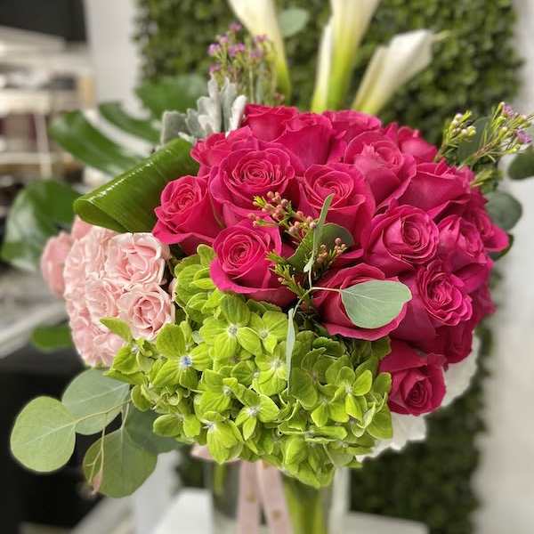 Bright pink roses, green hydrangeas, and pale pink blooms arranged in a clear glass vase with ribbon