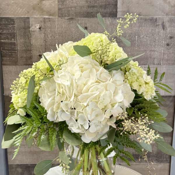 White and pale green hydrangea bouquet with lush foliage in a clear glass vase on a round white stand