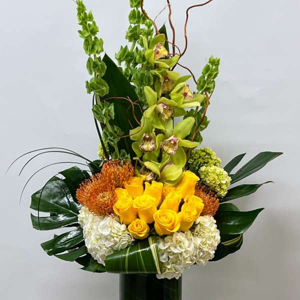 Tall floral arrangement with yellow roses, green orchids, and white hydrangeas in a glass vase