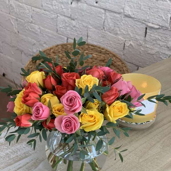 Round glass vase of red, pink, and yellow roses accented with eucalyptus on a light wood table.
