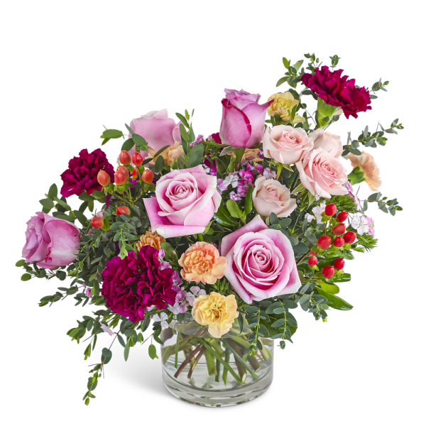 Short arrangement of pink roses, burgundy carnations, and peach blooms in a clear glass vase