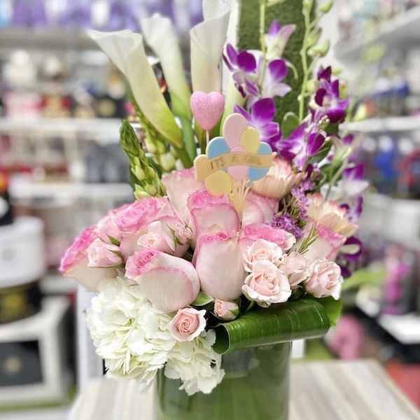 Tall arrangement of pink and white flowers with calla lilies in a green glass vase and an 'It's a Girl' pick